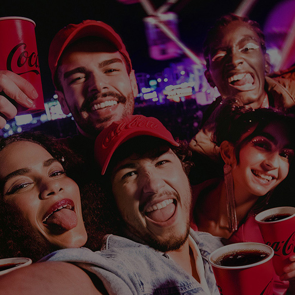 Grupo de jóvenes sonriendo y levantando vasos rojos de Coca-Cola en un ambiente festivo por la noche.