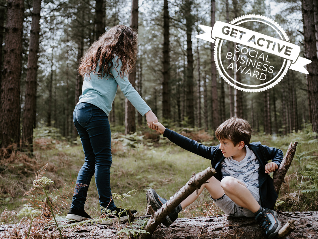 Ein Kind steht auf einem Baumstamm im Wald und reicht einem weiteren Kind das sitzt die Hand