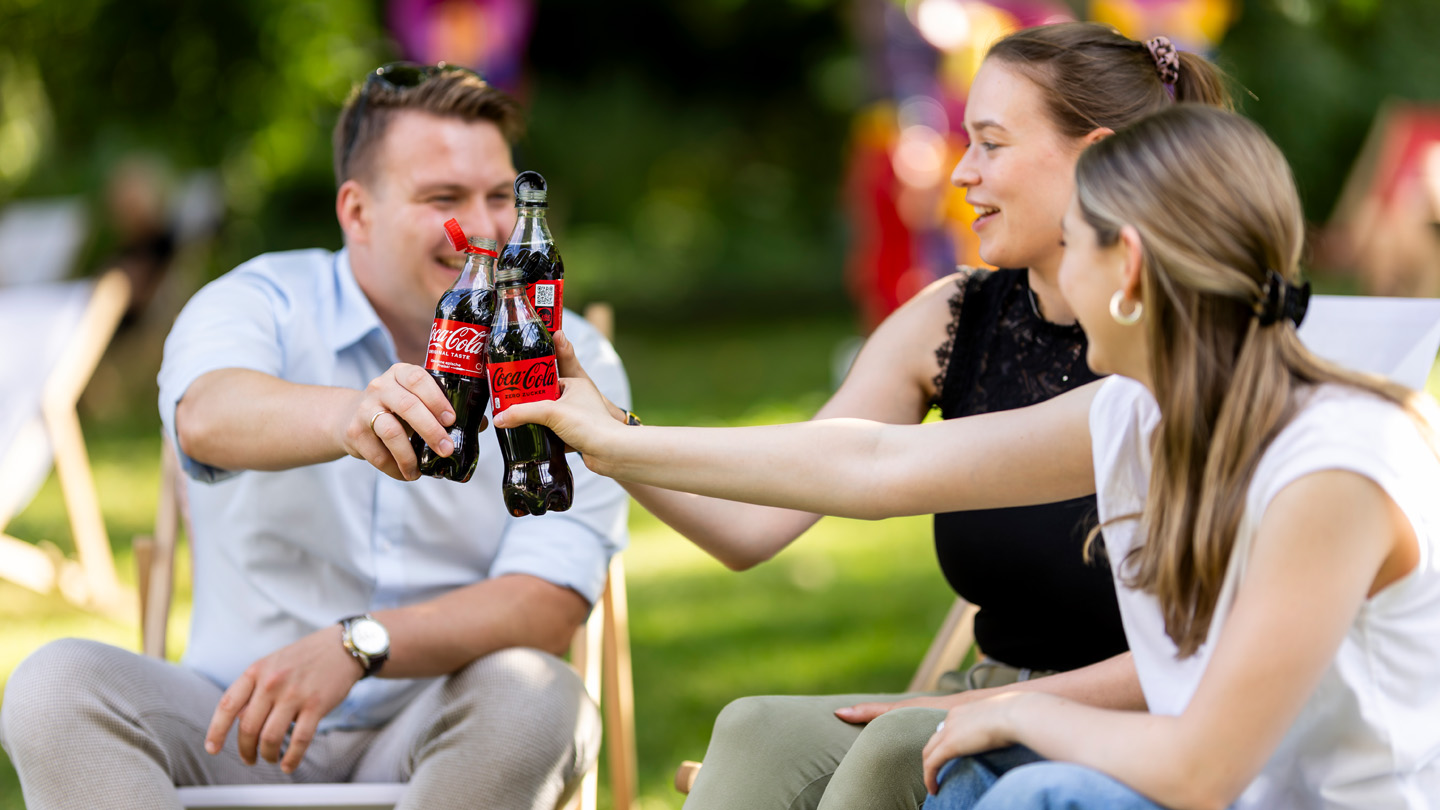 Drei Personen sitzen gemütlich und stoßen mit Coca-Cola Flaschen an