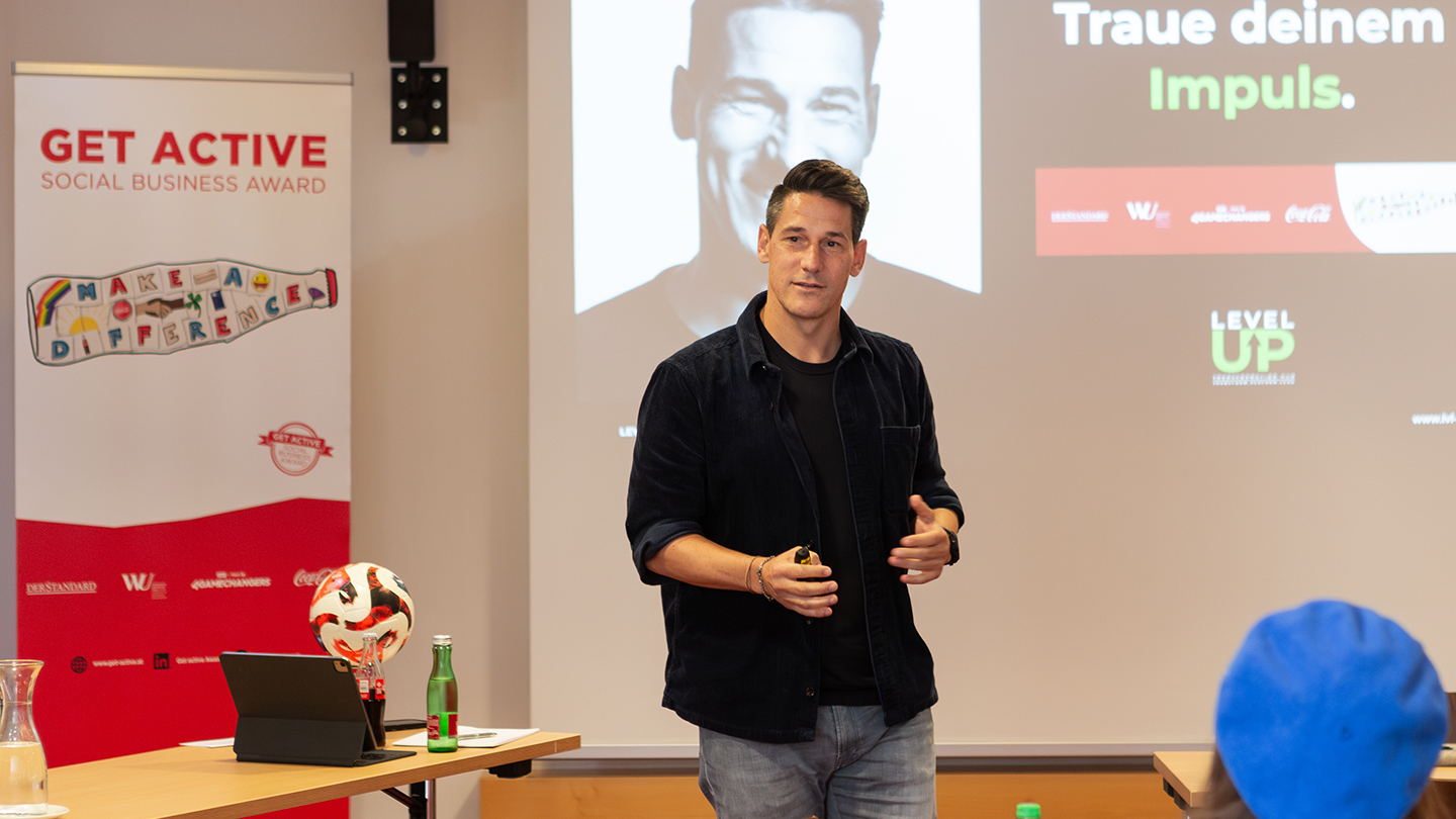 Ein Mann in dunkler Kleidung spricht auf einer Bühne beim Coaching-Wochenende. Neben ihm ein Banner mit „Get active Social Business Award“ sowie ein Tisch mit Fußball und Getränken. Auf der Leinwand ist sein Porträt zu sehen.