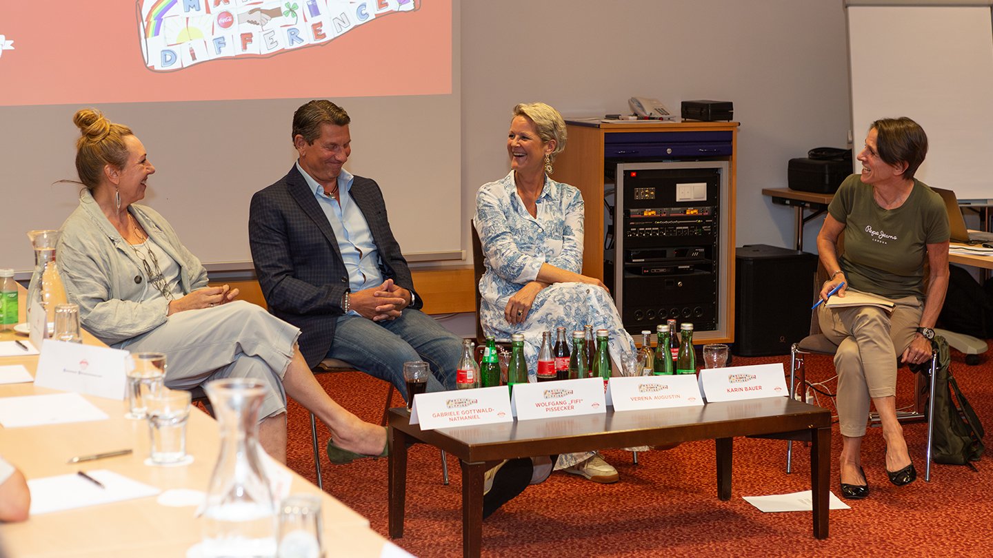 Vier Personen sitzen nebeneinander auf Stühlen und lachen im Gespräch. Vor ihnen ein Tisch mit Getränken, im Hintergrund eine Leinwand mit dem Slogan „Make a Difference“. Das Panel wird von Karin Bauer moderiert und bietet persönliche Geschichten aus dem Unternehmertum.