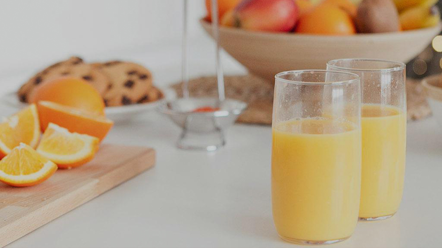 Deux verres remplis de jus d'orange frais sont posés sur une table blanche. À l'arrière-plan, une planche à découper avec des tranches d'orange, des biscuits, et un bol de fruits variés, comme des pommes et des kiwis.