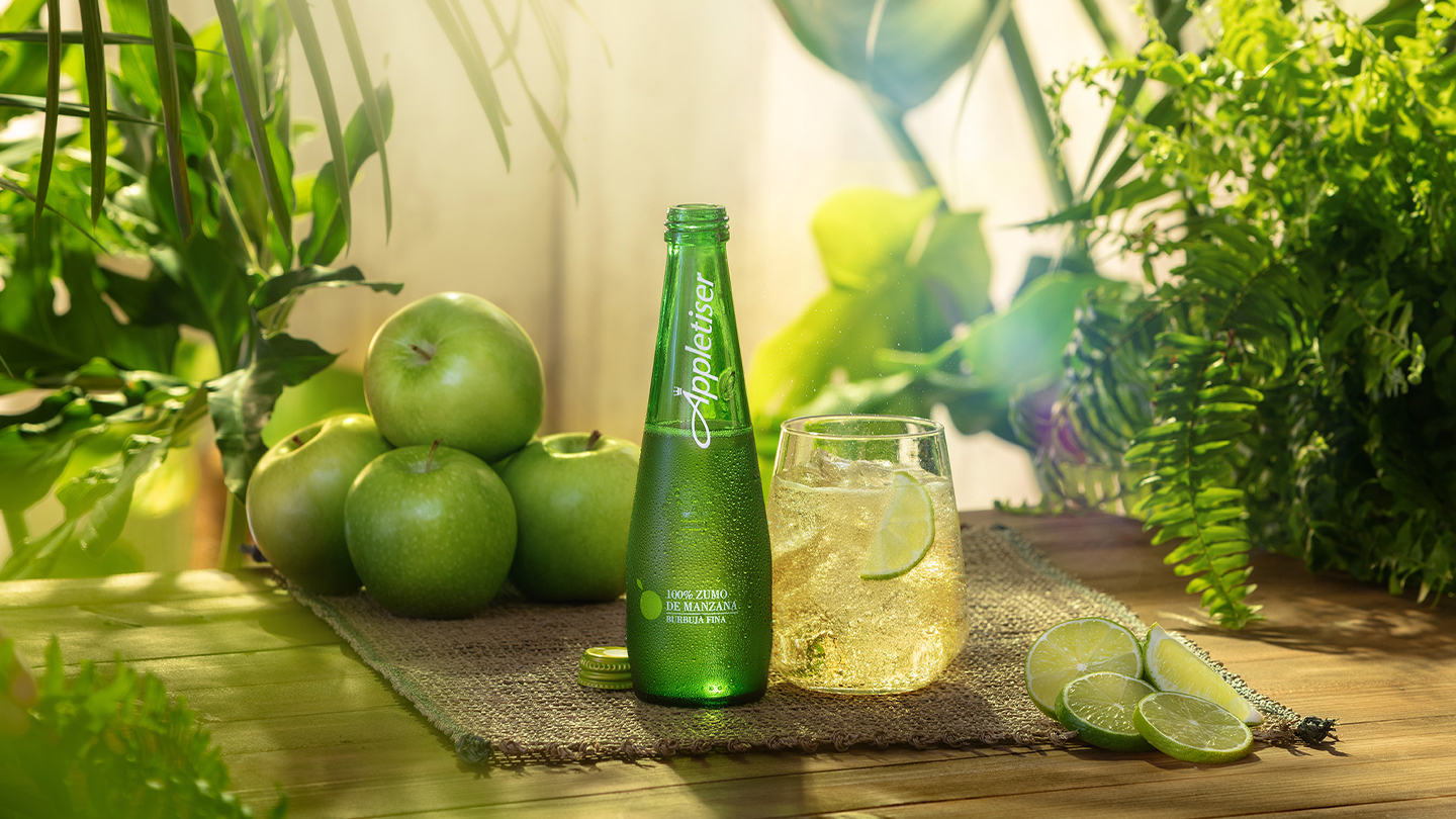Bouteille verte Appletiser, accompagnée d’un verre rempli de boisson pétillante, posés sur une table décorée de pommes vertes et tranches de citron vert, dans un décor lumineux abondant en plantes vertes.