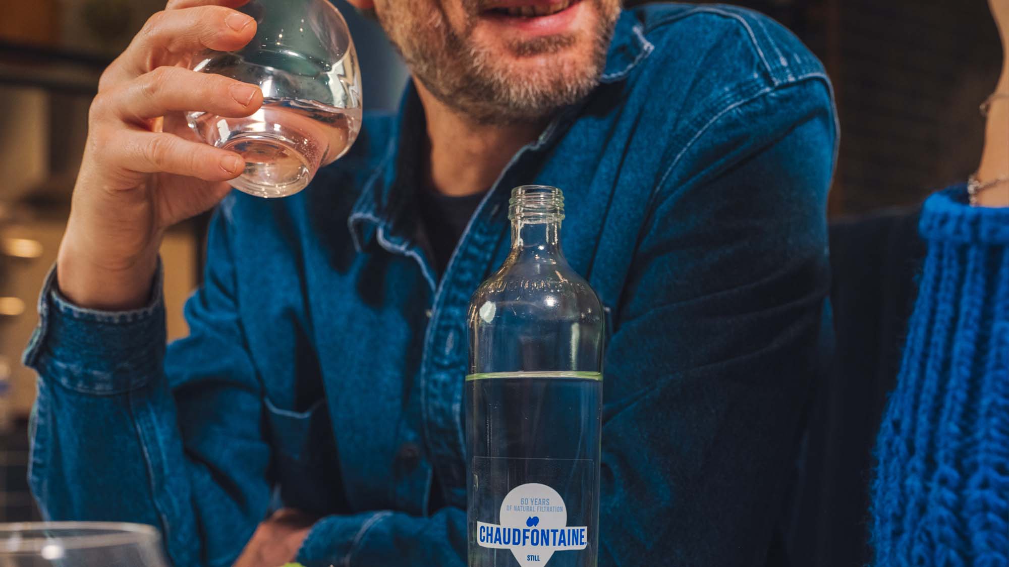 Gros plan sur un homme vêtu d’une chemise en jean, tenant un verre d’eau près de sa bouche, avec une bouteille en verre Chaudfontaine posée sur la table. On aperçoit une autre personne portant un pull bleu à droite de l’image.