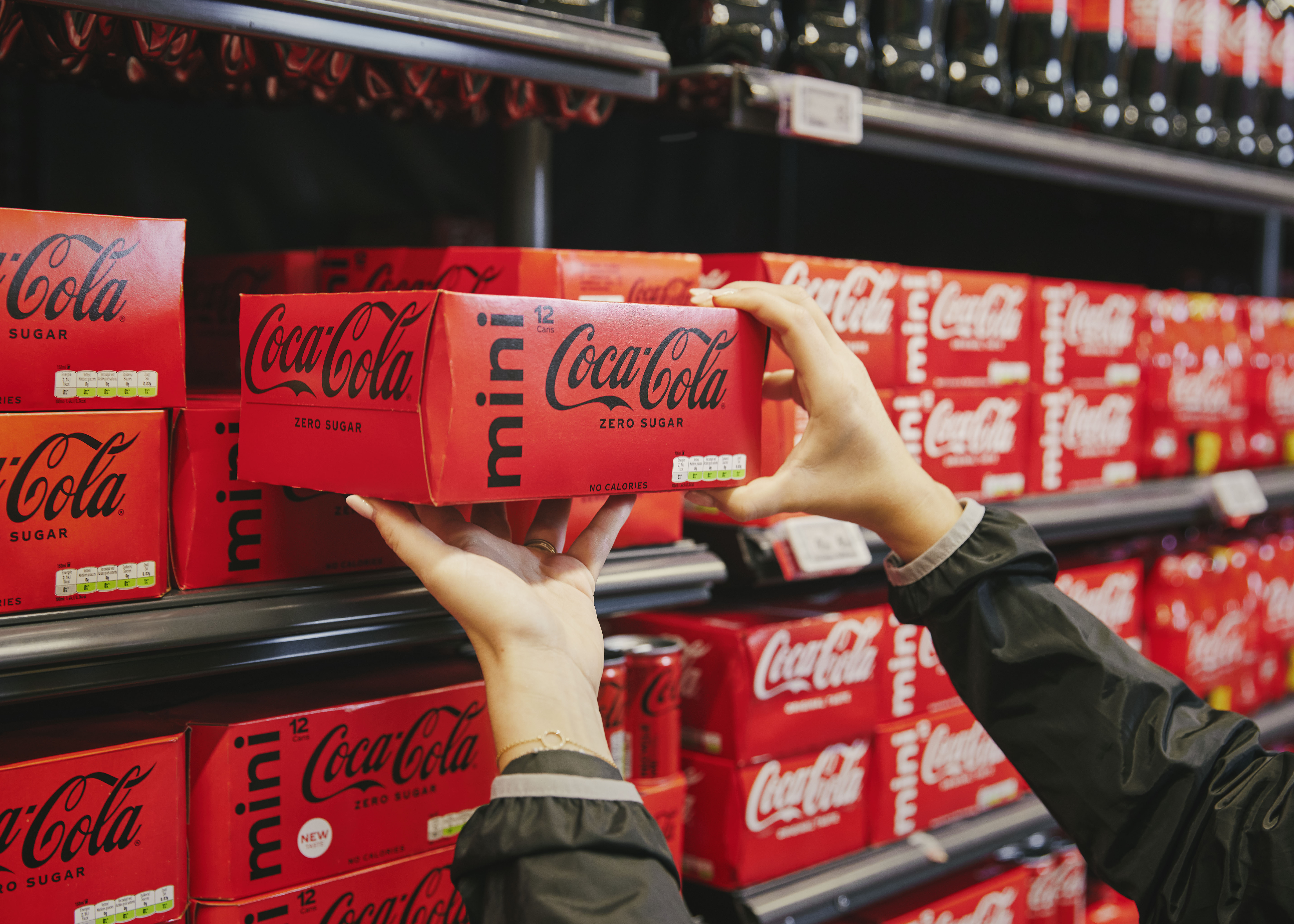 Foto met handen die een pak Coca‑Cola Zero Sugar mini in een supermarkt schap plaatsen, omringd door merkproducten.