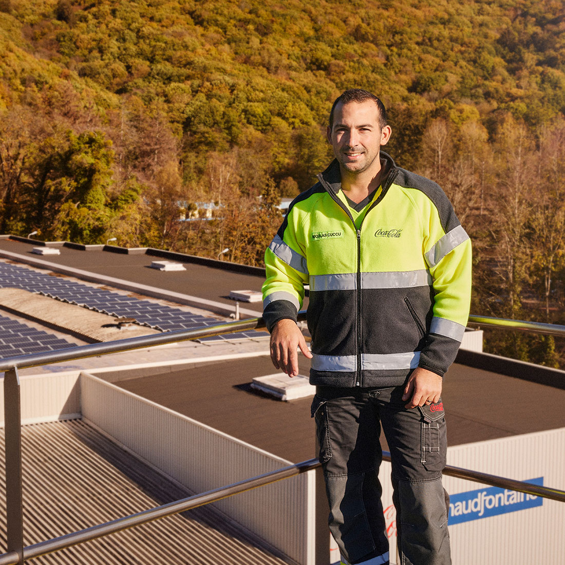 Foto met een werknemer in hoogzichtbare veiligheidskleding op het dak van de Chaudfontaine‑fabriek in België, met zonnepanelen en een beboste heuvel op de achtergrond.
