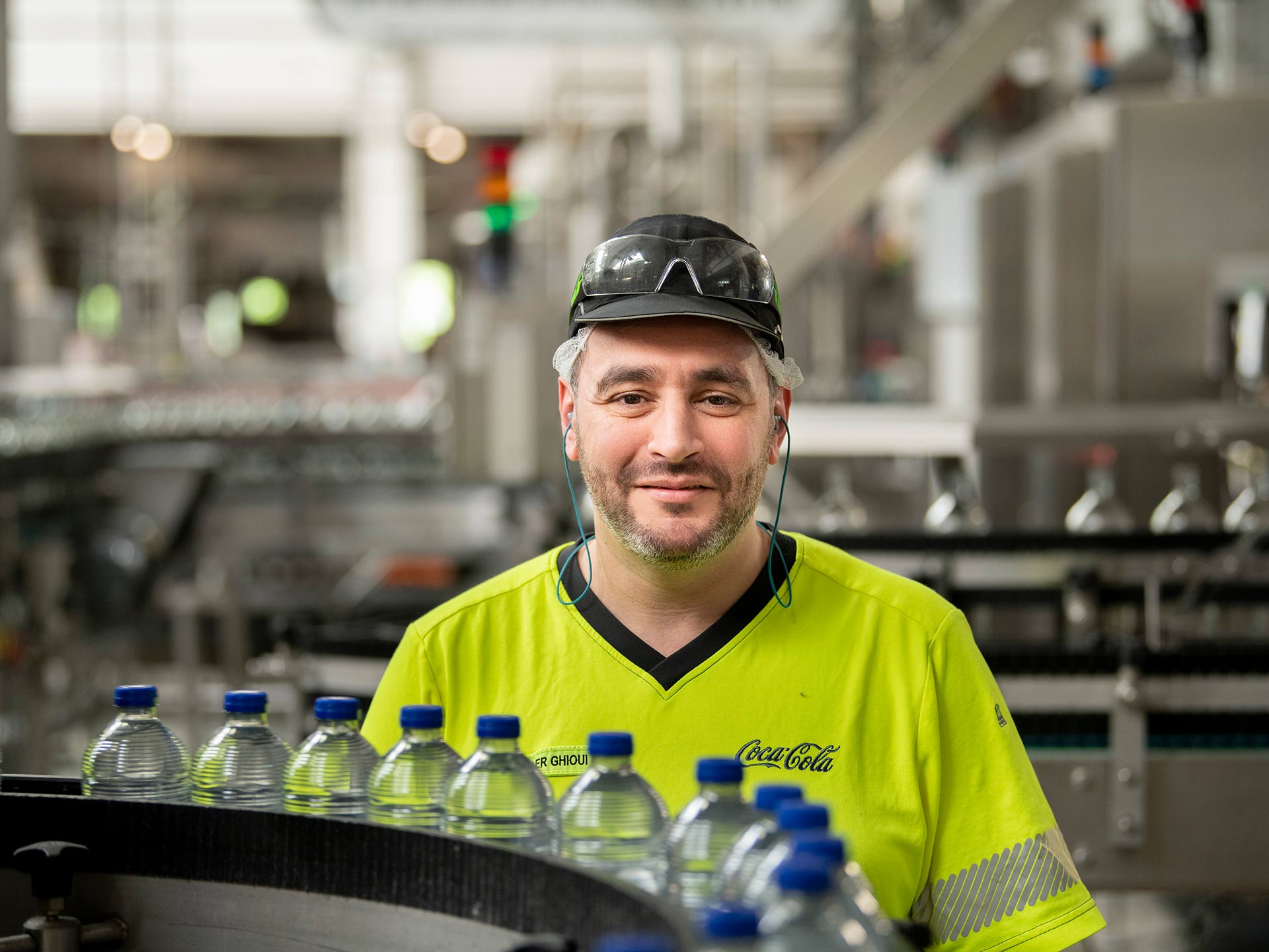 Photo avec un employé en tenue de sécurité haute visibilité devant une chaîne de production de bouteilles d’eau.