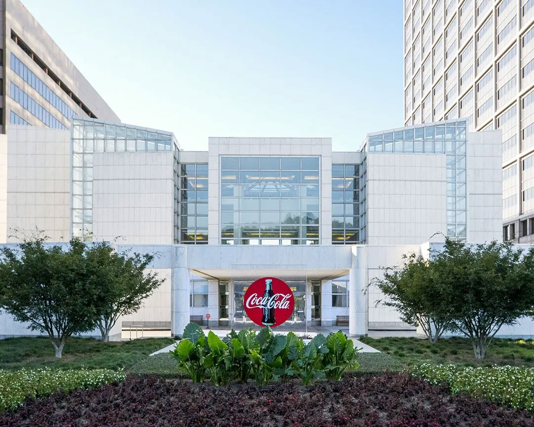 Foto met het moderne hoofdkantoor van Coca‑Cola Atlanta, witte glazen gevel, rood logo van het merk bij de ingang, omringd door groen en bomen.