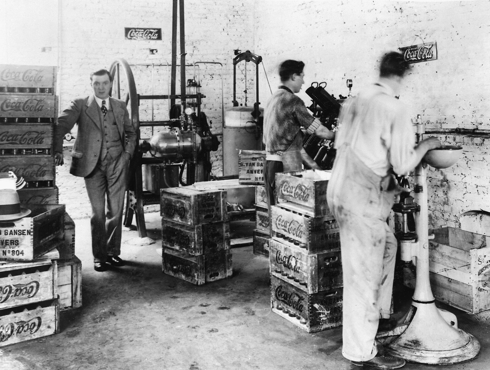 Embouteillage à Anvers par Gustave Van Gansen : Photo avec une ancienne usine Coca‑Cola où des personnes travaillent parmi des caisses en bois et des machines de production.