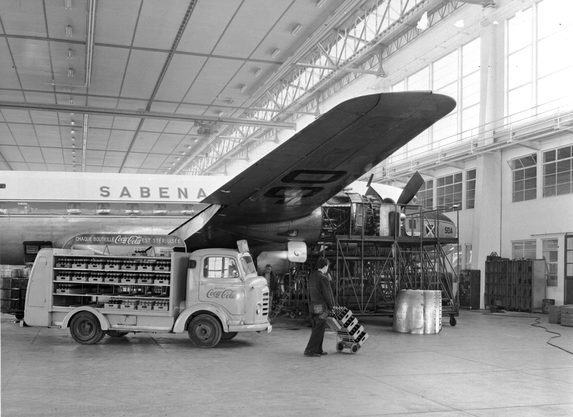 Photo avec un avion de la compagnie Sabena dans un hangar, à côté d’un camion Coca‑Cola livré avec des caisses de bouteilles.