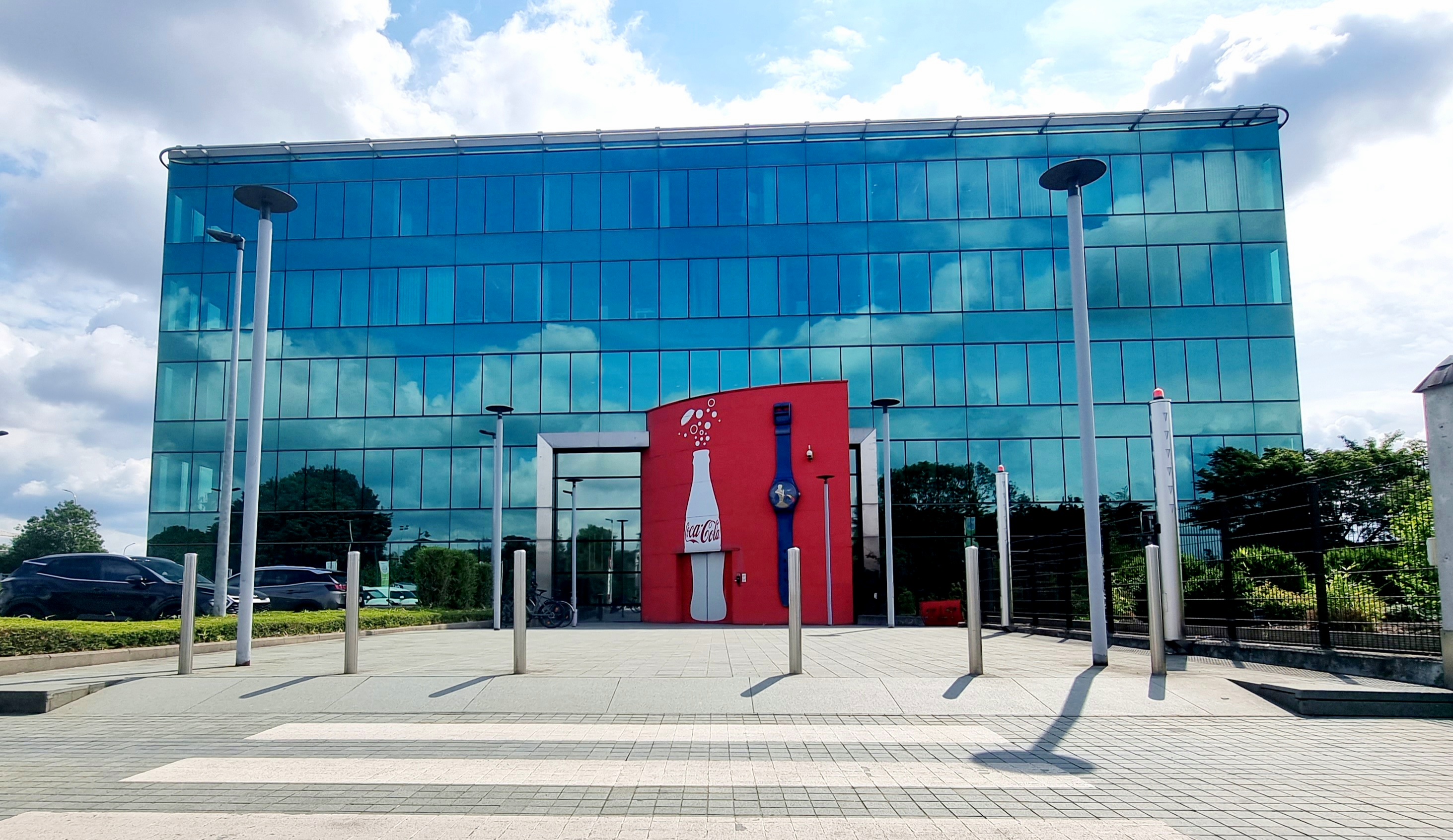 Foto met het hoofdkantoor van Coca‑Cola in België, modern gebouw met een blauwe glazen gevel, ingang versierd met een rode muur met een witte Coca‑Cola fles en een groot decoratief horloge.