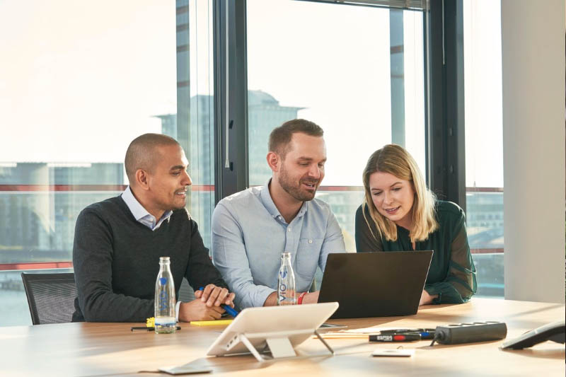 Foto met drie mensen rond een vergadertafel, werkend op een laptop, met Coca‑Cola waterflessen op tafel en uitzicht op de stad door de ramen.