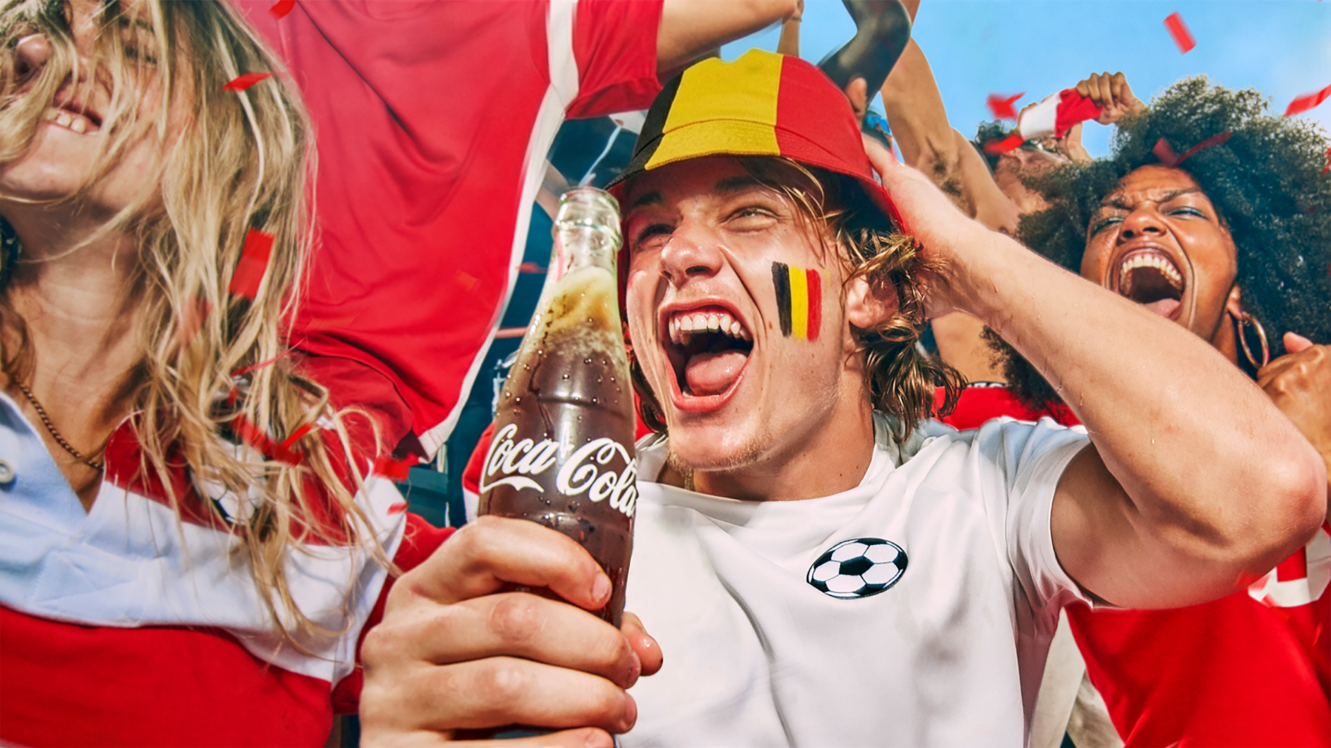 Un groupe de supporters de football en tenues rouge et blanc célèbre avec des confettis ; au premier plan, une personne tient une bouteille de Coca‑Cola.
