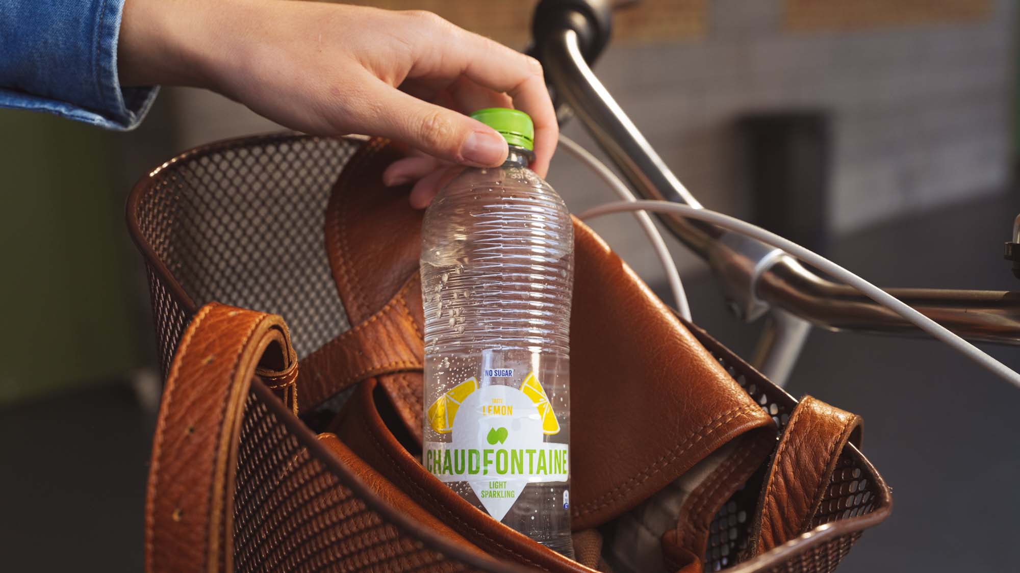 Hand haalt een plastic fles Chaudfontaine Light Sparkling met citroensmaak, zonder suiker, met groene dop, uit een bruine leren tas in een fietsbmand.