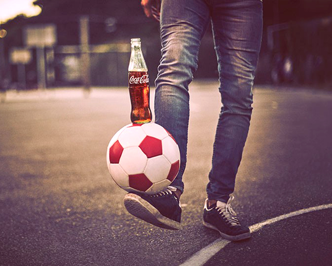 De foto toont een persoon van wie alleen de benen zichtbaar zijn, staand op een sportveld, met een voetbal in evenwicht op de voet. Op de bal staat een glazen fles Coca-Cola.