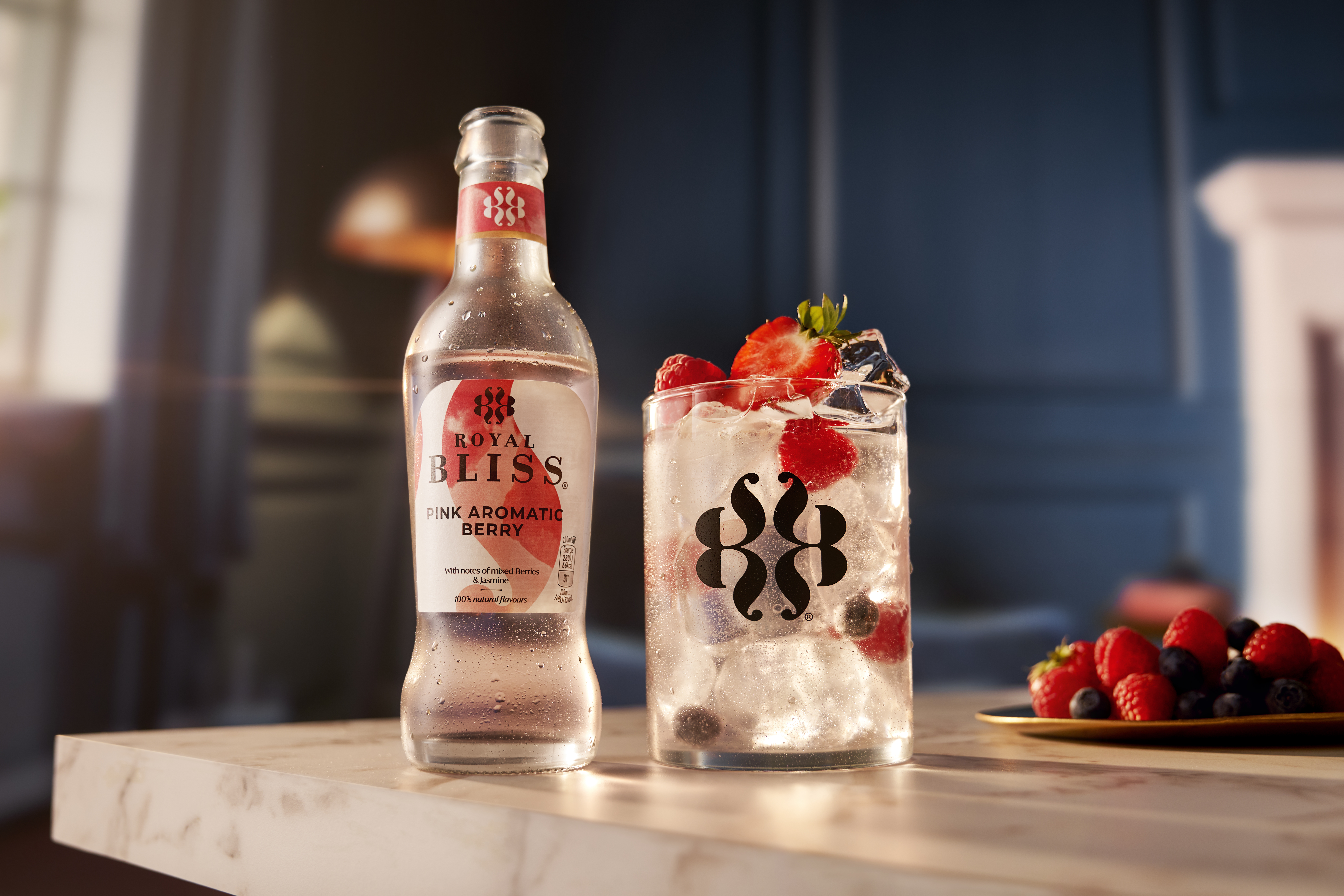 Fles “Royal Bliss – Pink Aromatic Berry” met lichtroze inhoud op een werkblad naast een glas met ijsklontjes en hele rode en blauwe bessen in de bruisende drank. Op de achtergrond een warme setting met een bord vol rood fruit.