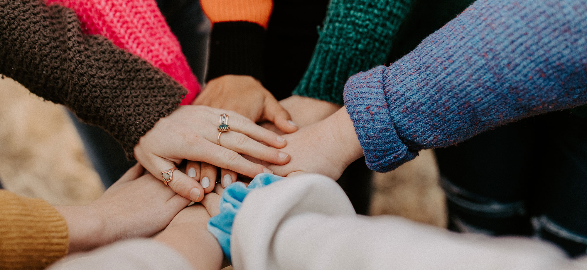 Des mains de plusieurs personnes, portant des pulls colorés, sont posées les unes sur les autres en signe d’unité et de solidarité. Certaines mains portent des bagues.
