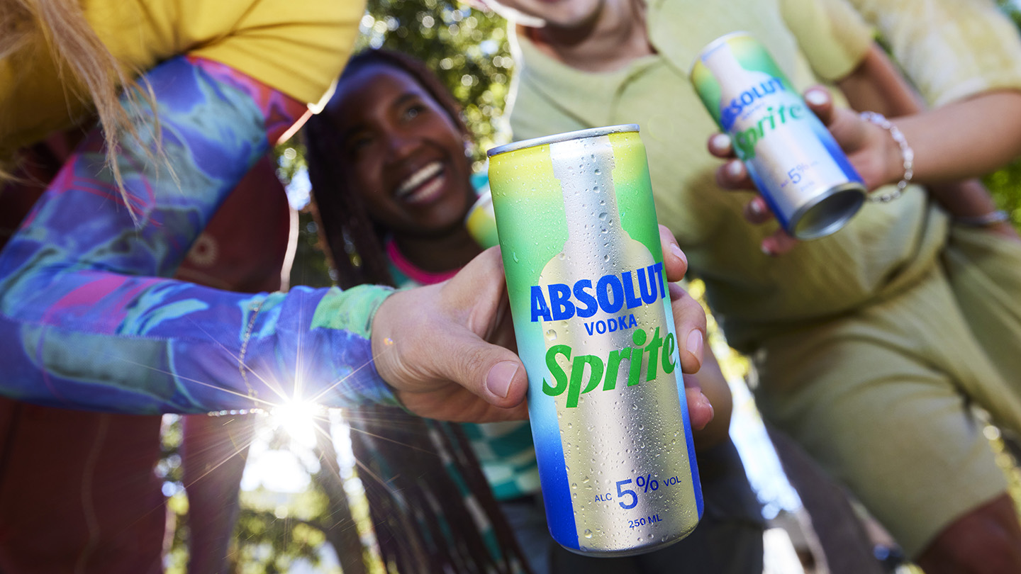 Lata de Absolut Vodka Sprite em primeiro plano, com uma mão segurando-a e uma pessoa rindo ao fundo
