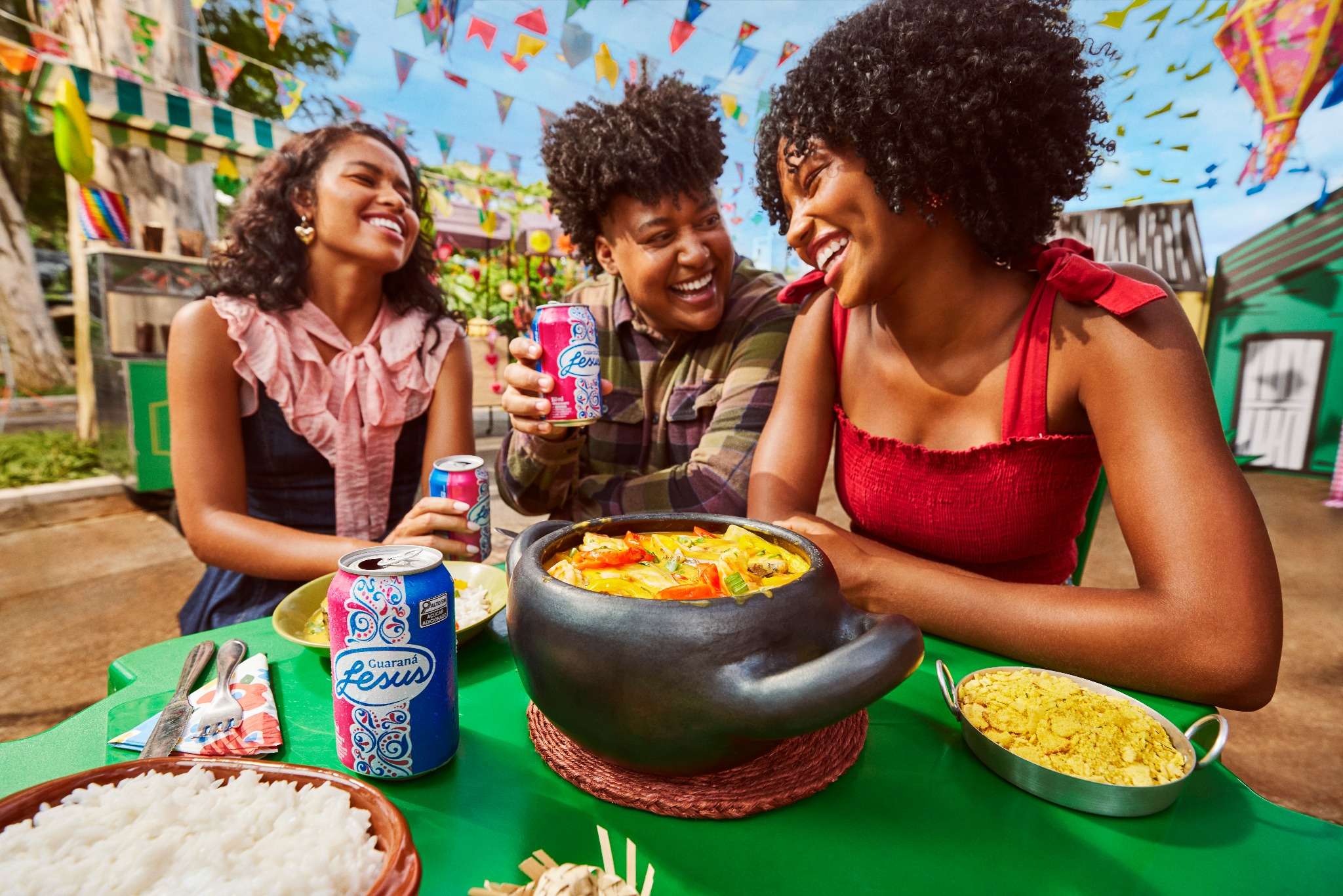 Três amigos desfrutam de Guaraná Jesus e pratos típicos em uma animada festa.