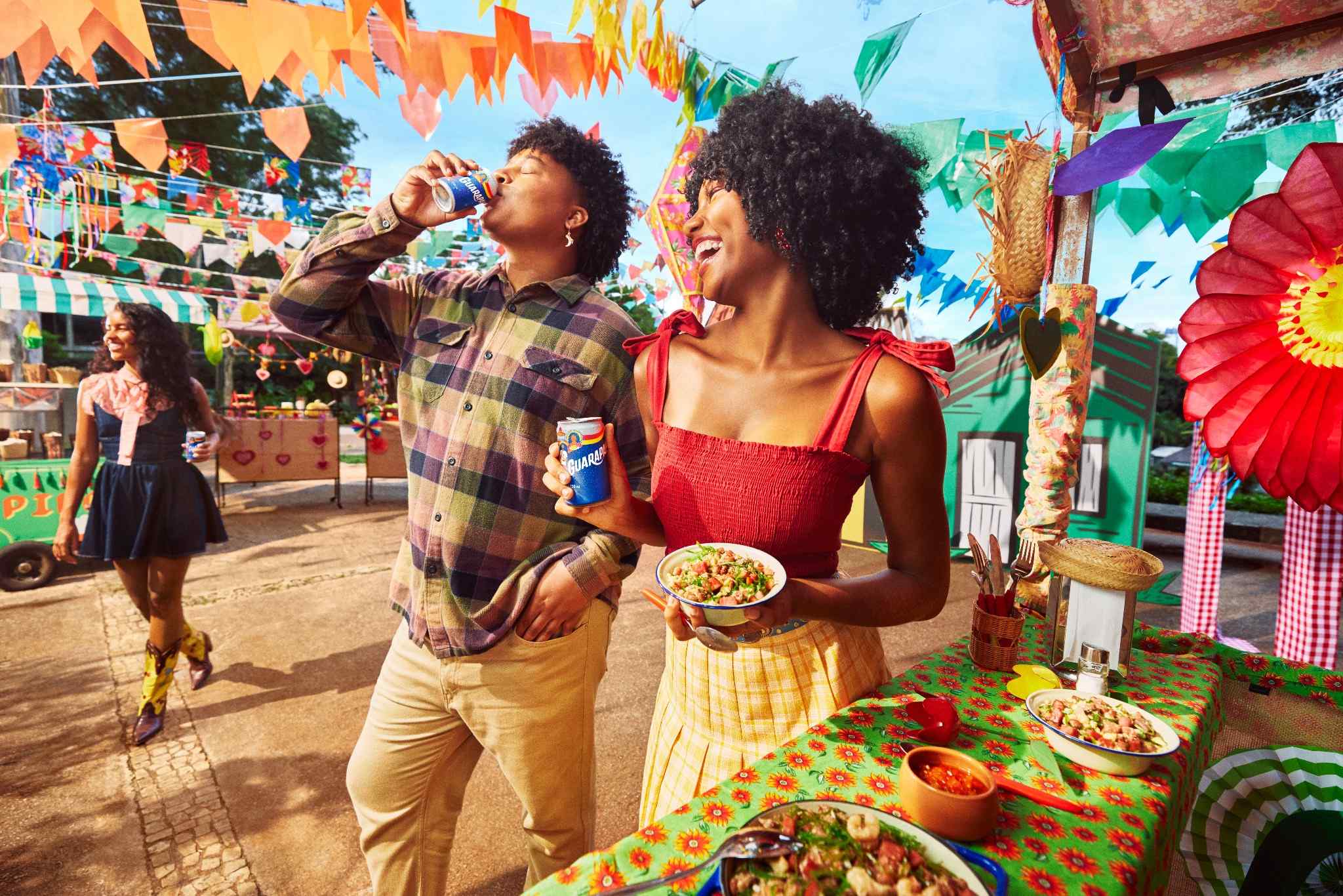 Amigos sorrindo em uma Festa, com Guarapan, mesa farta de comidas típicas, bandeirinhas e decoração colorida.