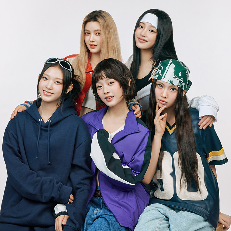 NewJeans vestindo roupas casuais e estilosas, incluindo moletons, jaquetas esportivas e bandanas. Eles estão sorrindo e posando juntos em frente a um fundo branco, transmitindo um espírito jovem e descontraído.