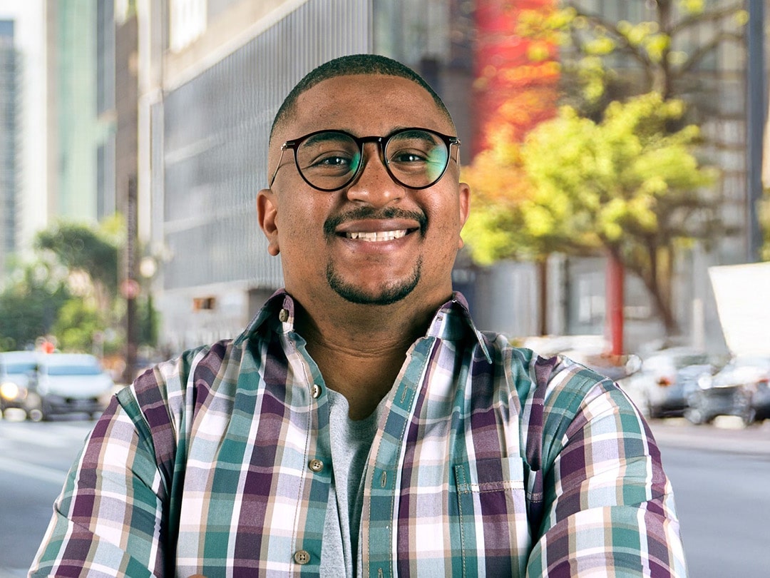 Um homem negro de óculos e camisa xadrez sorrindo em uma rua movimentada da cidade.