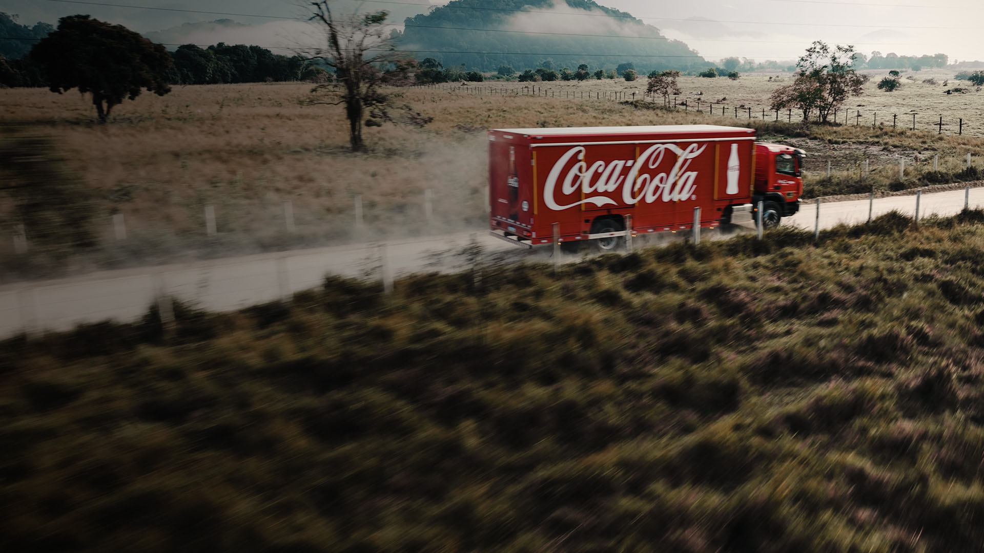Caminhão vermelho da Coca-Cola em movimento em uma estrada de terra, com paisagem rural e montanhas ao fundo.
