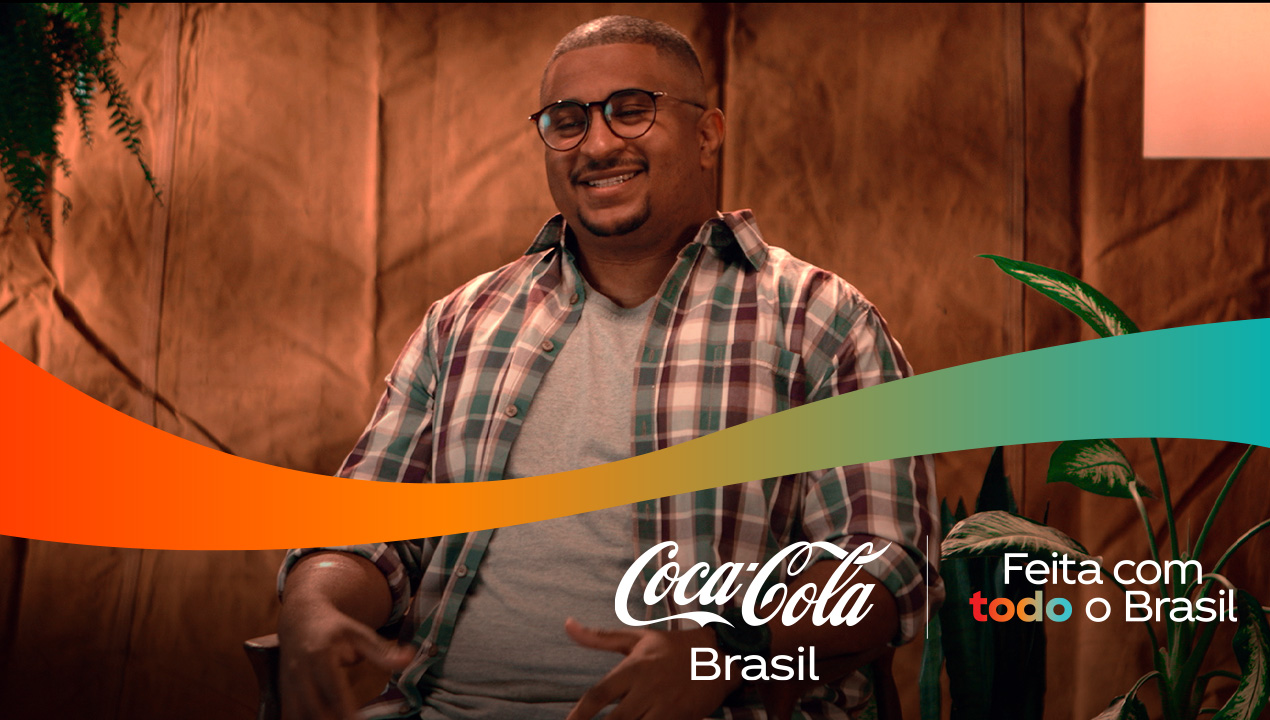 Um homem sorrindo, usando óculos e camisa xadrez, com o logo da Coca-Cola Brasil e a frase "Feita com todo o Brasil" sobre uma faixa colorida.