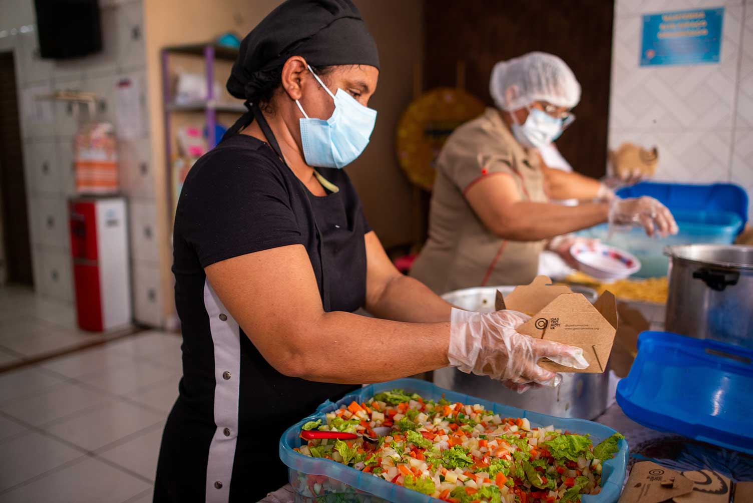 As pessoas que trabalham na cozinha usam máscaras, luvas e toucas de cabelo. Na mesa há comida e panelas.
