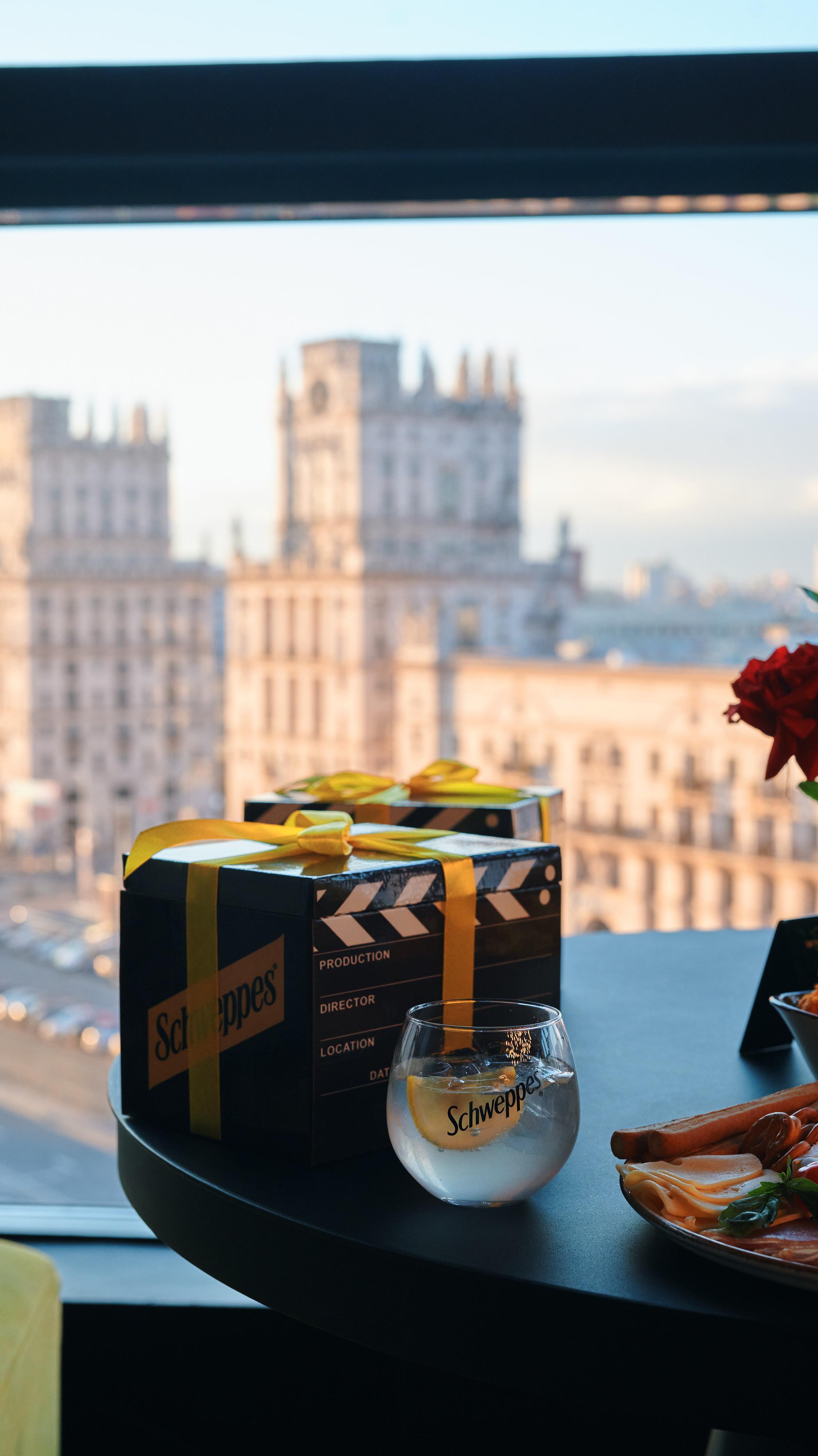 Падорачныя каробкі Schweppes і шклянка з напоем на стале з выглядам на горад