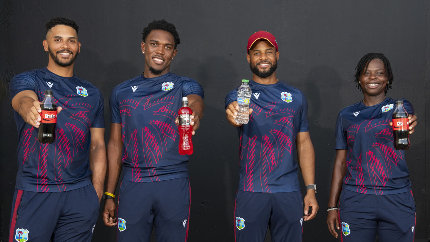 Cuatro jugadores de West Indies Cricket, dos hombres y dos mujeres, sonríen mientras sostienen botellas frente a un fondo oscuro.