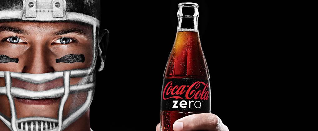 Mann mit aufgemaltem Rugbyhelm hält eine Coca-Cola Zero Zucker Flasche in der Hand auf schwarzem Hintergrund