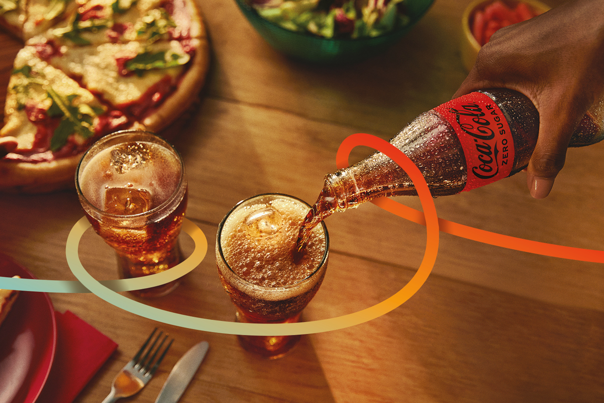 Eine Hand hält eine Coca-Cola Zero Sugar Flasche und füllt ein Coca-Cola Glas auf einem mit Pizza und Salat gedeckten Tisch