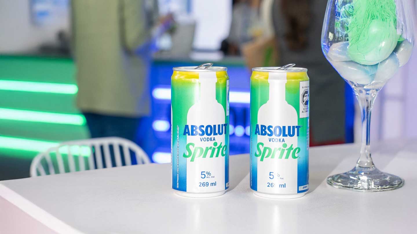  Dos latas de Absolut Sprite y una copa con un elemento verde en una mesa blanca, con un fondo borroso de luces de neón.