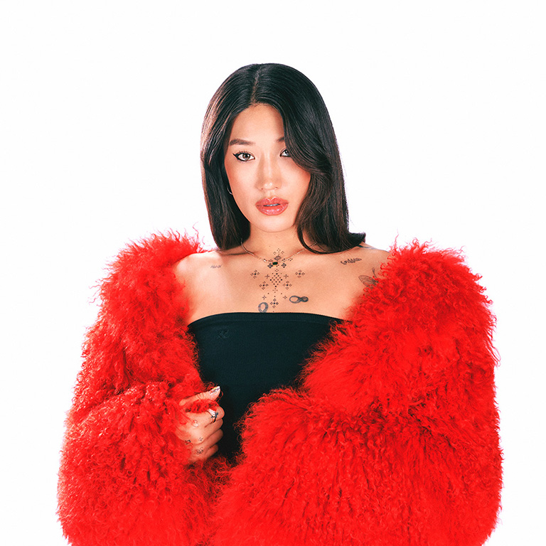 Peggy Gou, con el pelo largo y oscuro, lleva un top negro sin tirantes y un gran abrigo rojo. Tiene varios tatuajes en el pecho y el cuello, incluyendo estrellas y diseños celestiales. Está mirando directamente a la cámara con una expresión confiada. El fondo es blanco, destacando su vibrante atuendo.