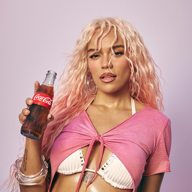 Karol G de cabello largo rubio sosteniendo una botella de Coca-Cola. Lleva una remera rosa atada al frente y un bikini blanco debajo.