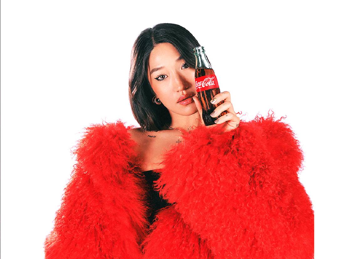 Peggy Gou sosteniendo una botella de Coca-Cola mientras viste una chaqueta de piel roja, en un fondo blanco.