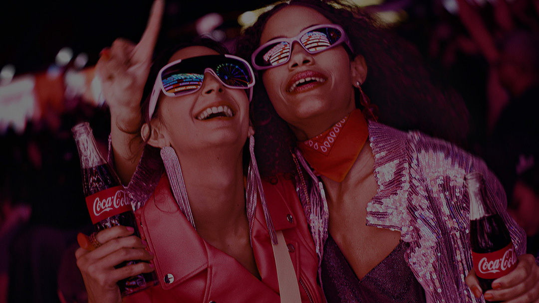 Dos amigas sostenido una botella de Coca-Cola en la mano sonriendo en un ambiente festivo.