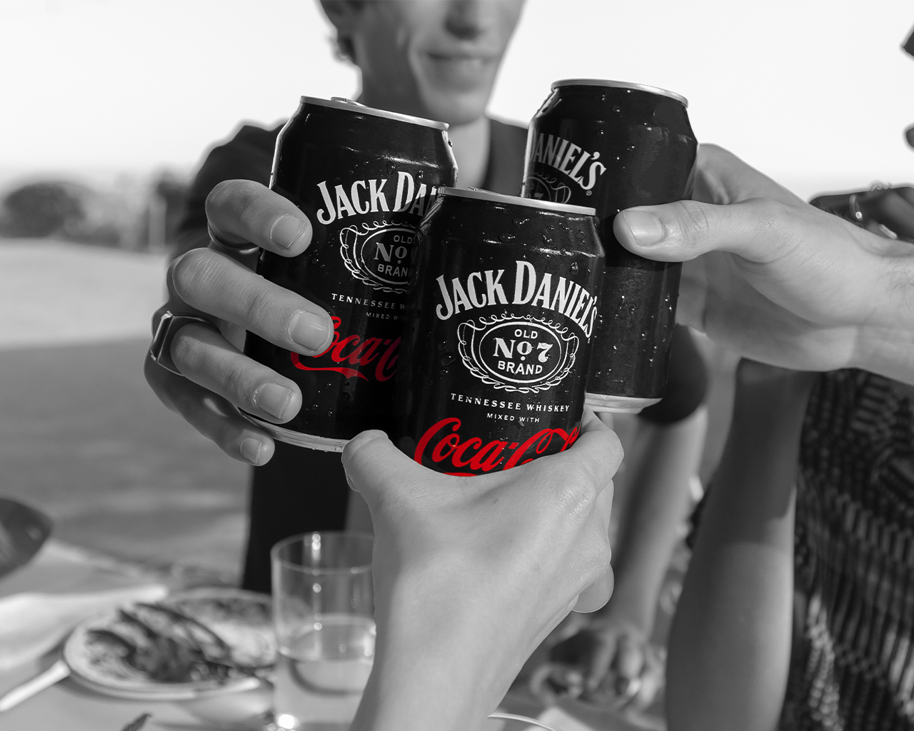 Tři ruce si připíjejí s plechovkami Jack Daniel's a Coca-Cola na černobílém pozadí.
