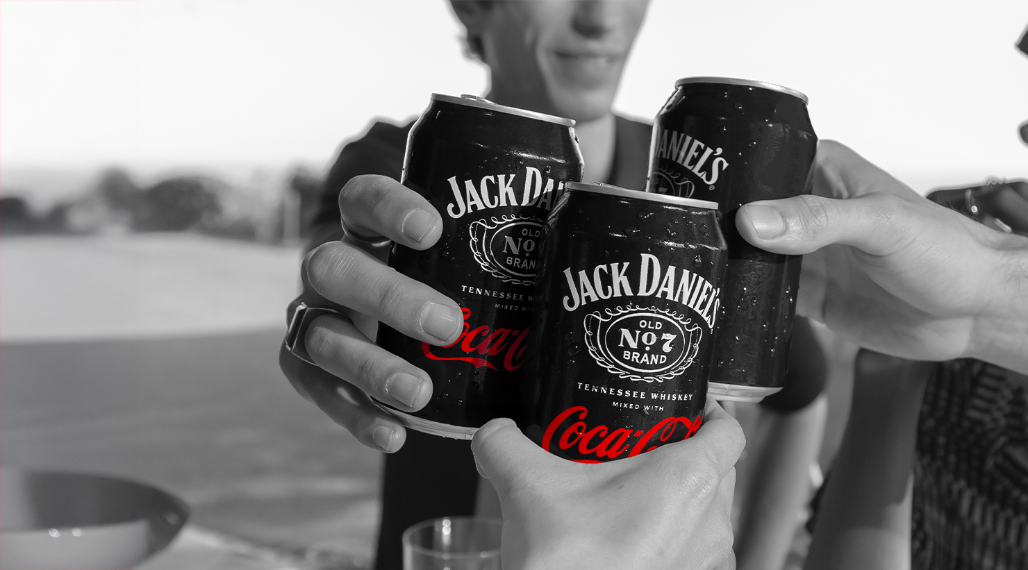 Tři ruce si připíjejí s plechovkami Jack Daniel's a Coca-Cola na černobílém pozadí.