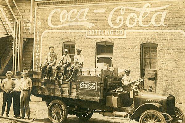 Ajalooline foto Coca‑Cola veokist, millel istuvad töömehed, taustal Coca‑Cola villimistehas.
