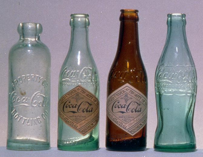 Neli ajaloolist Coca‑Cola klaaspudelit, erinevates värvitoonides ja kujundustes, pärit 19.–21. sajandist.