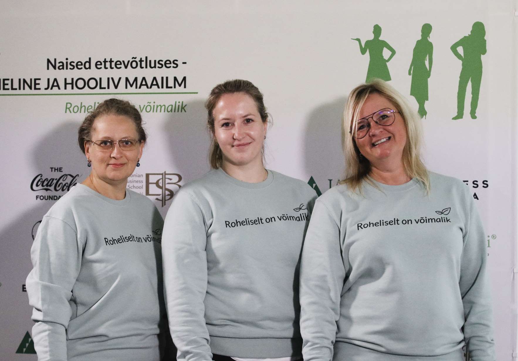 Kolm inimest hallides “Roheliselt on võimalik” pusades naiste ettevõtluse bänneri ees.
