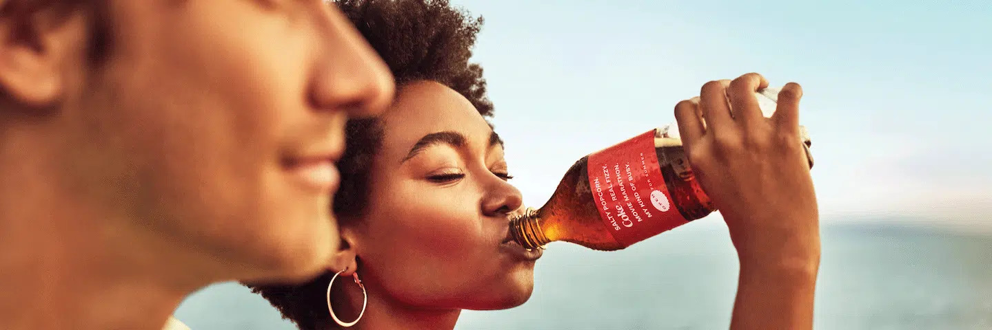 nimene hoiab ja joob Coca-Cola pudelist, taustal merel vaade; noorte võimestamise teemal.