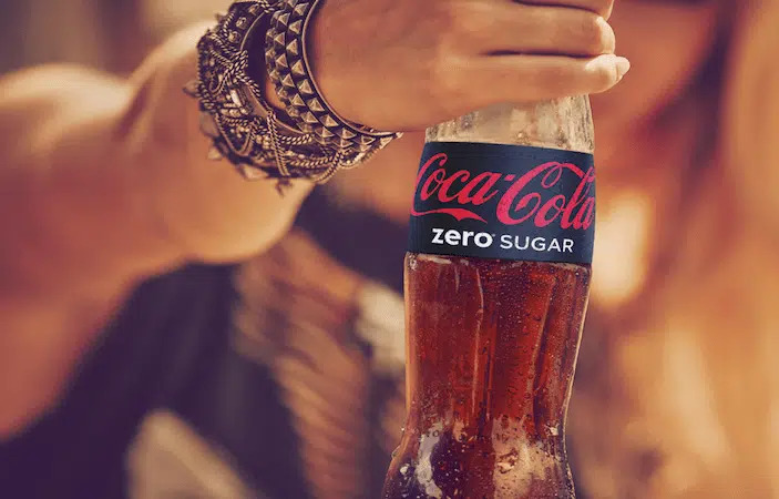 Käsi hoiab Coca-Cola Zero Sugar klaaspudelit, käevarel mitmed metallist ja nahast käevõrud.