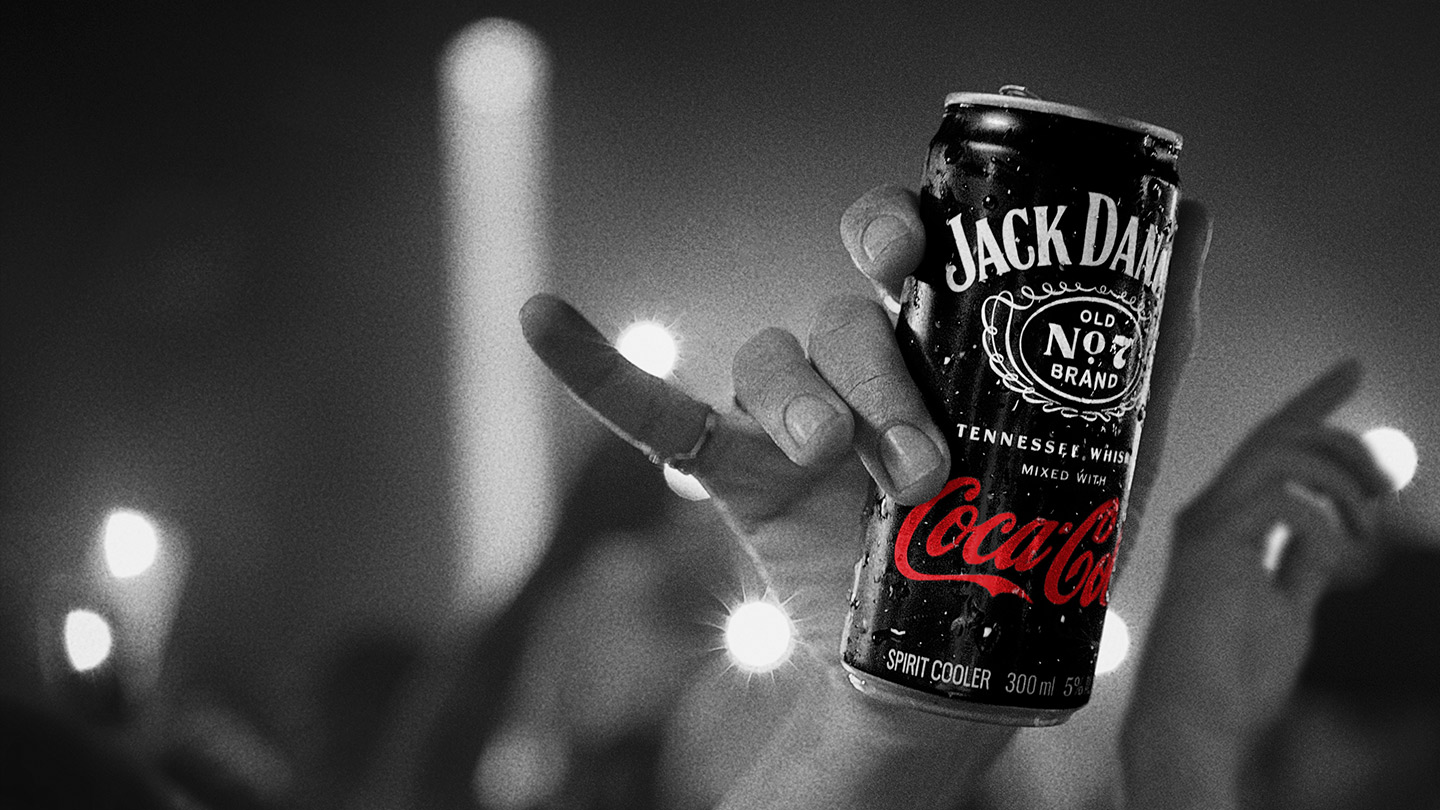 Image d'une main, dans une ambiance de fête ou de concert, brandissant une canette noire de Jack Daniel's & Coca-Cola. En arrière-plan, des lumières et des silhouettes d'autres mains sont visibles.