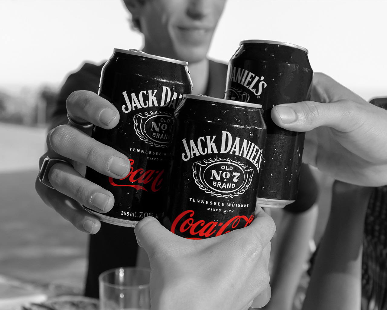 Tri ruke drže i nazdravljaju limenkama „Jack Daniel’s & Coca-Cola“ u crno-beloj fotografiji sa naglašenim crvenim logoom.