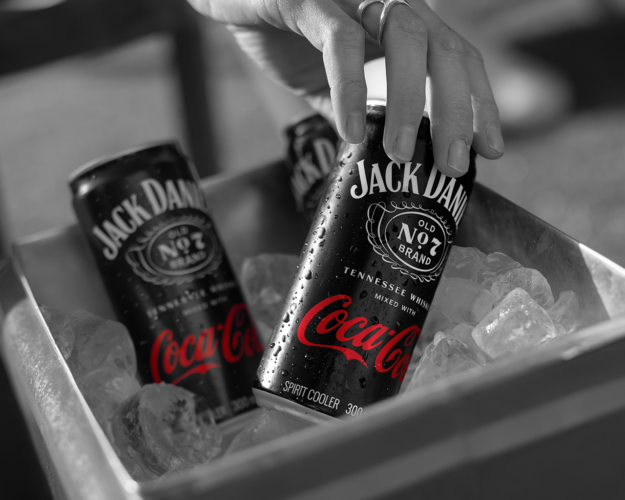 Une main saisit une canette Jack Daniel’s & Coca‑Cola posée dans un bac rempli de glace, avec une autre canette visible en arrière‑plan. L’image est en noir et blanc, la canette mise en avant évoquant la fraîcheur et le goût.