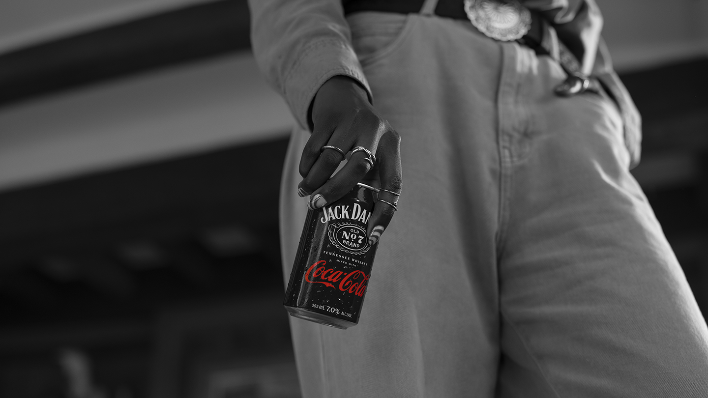 Osoba drži crnu limenku „Jack Daniel’s & Coca-Cola“ uz bok, sa vidljivim crvenim logoom na crno-beloj pozadini.