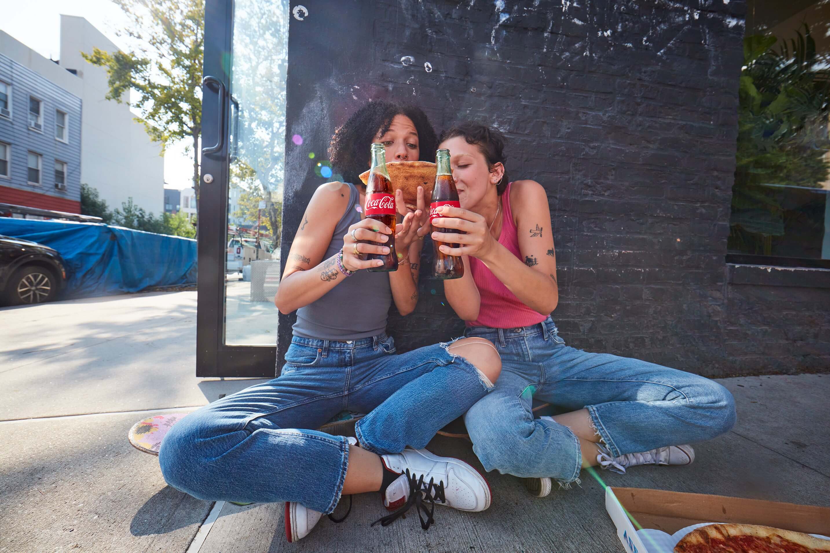 Photo avec deux personnes assises sur un trottoir, tenant des bouteilles Coca‑Cola et partageant une part de pizza